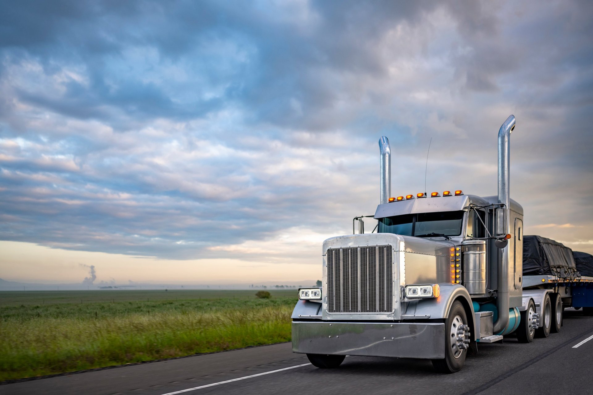 Gray classic big rig American semi truck with turned on lights and chrome exhaust pipes transporting covered cargo on flat bed semi trailer driving on the twilight highway road with stormy clouds sky