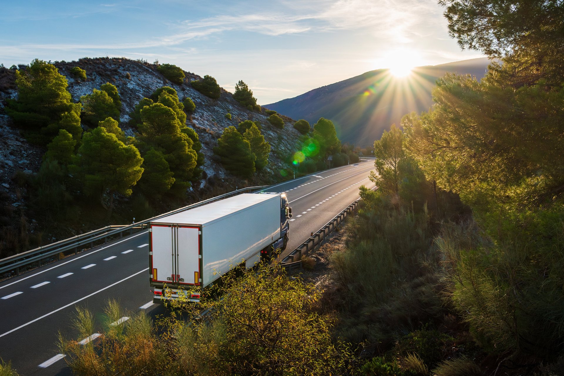 Refrigerated truck driving along mountain road with sunlight breaking over horizon