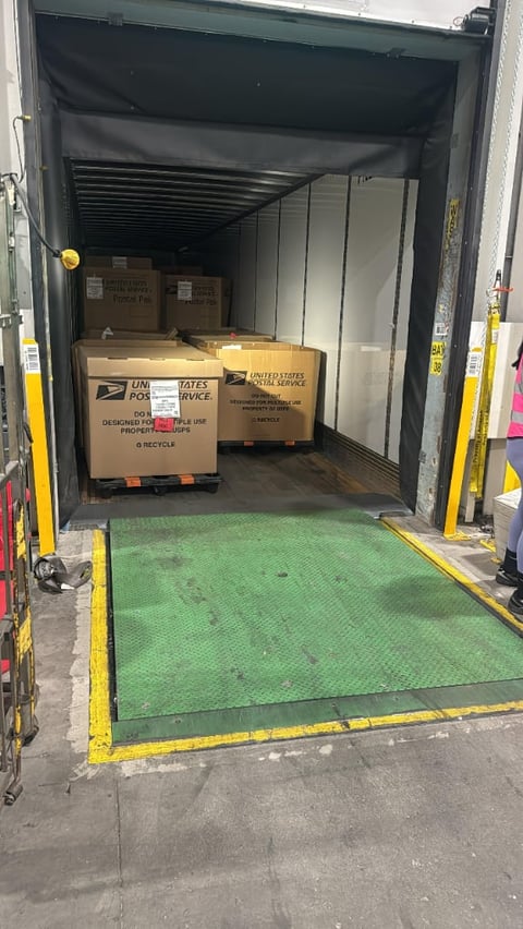 Open delivery truck with cardboard boxes labeled United States Postal Service loaded inside, green loading ramp visible in foreground