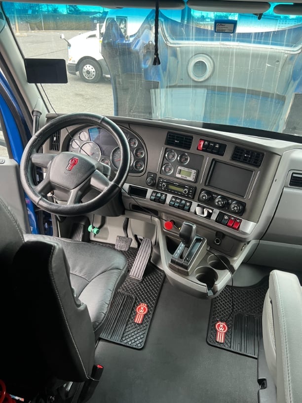 Interior of semi-truck cab showing steering wheel, dashboard controls, and windshield view of parked trucks